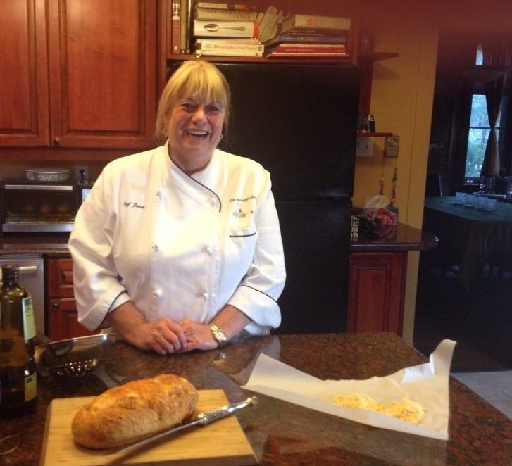 Janet Craig in her kitchen, wearing a chef's outfit in front of a marble counter with a loaf of bread on top, a glass of red wince and olive oil